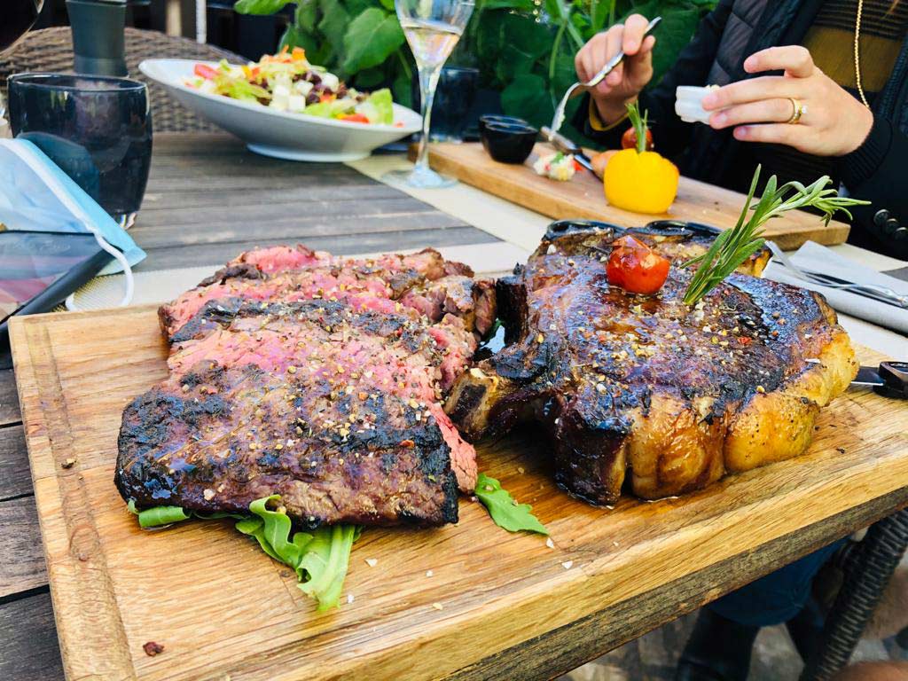 Zwei gegrillte Steaks, eines in Scheiben geschnitten, auf einem Holzbrett mit Rosmarin garniert. Im Hintergrund ein Salat und eine Person, die isst.