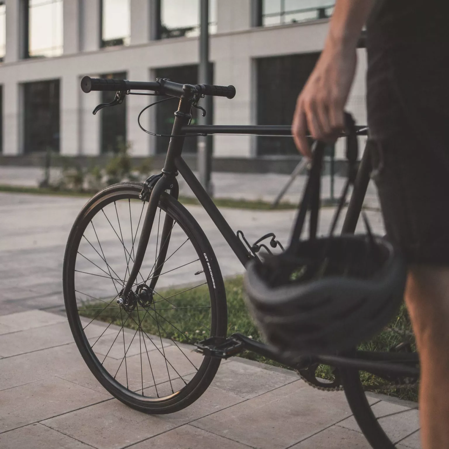 Eine Person steht neben einem schwarzen Fahrrad und hält einen Helm in der Hand. Im Hintergrund ist ein modernes Gebäude bei schwachem Licht zu sehen. Die Szene wirkt ruhig und urban.