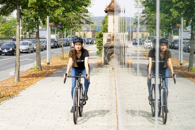 Eine Frau mit Helm sitzt lächelnd auf einem Fahrrad, umgeben von einem modernen städtischen Umfeld mit Pflanzen im Vordergrund.