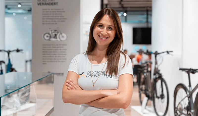 Eine Frau im T-Shirt der Deutschen Dienstrad steht mit verschränkten Armen in einem modernen Raum mit Fahrrädern im Hintergrund und lächelt in die Kamera.