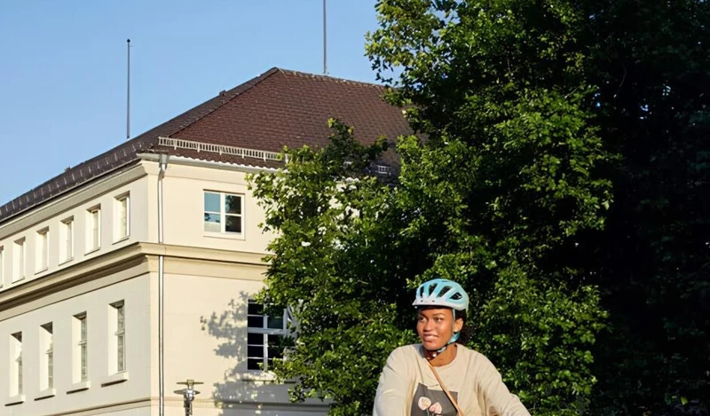 Zwei Personen fahren auf E-Bikes durch eine sonnige, städtische Umgebung. Beide tragen Helme, und im Hintergrund ist ein großes Gebäude mit klassischen Säulen zu sehen.