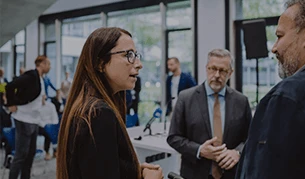 Eine Frau mit Brille unterhält sich mit zwei Männern in einem modernen Konferenzraum. Einer der Männer trägt einen Anzug und Krawatte, während der andere lässiger gekleidet ist. Im Hintergrund sind weitere Personen und große Fenster zu sehen.