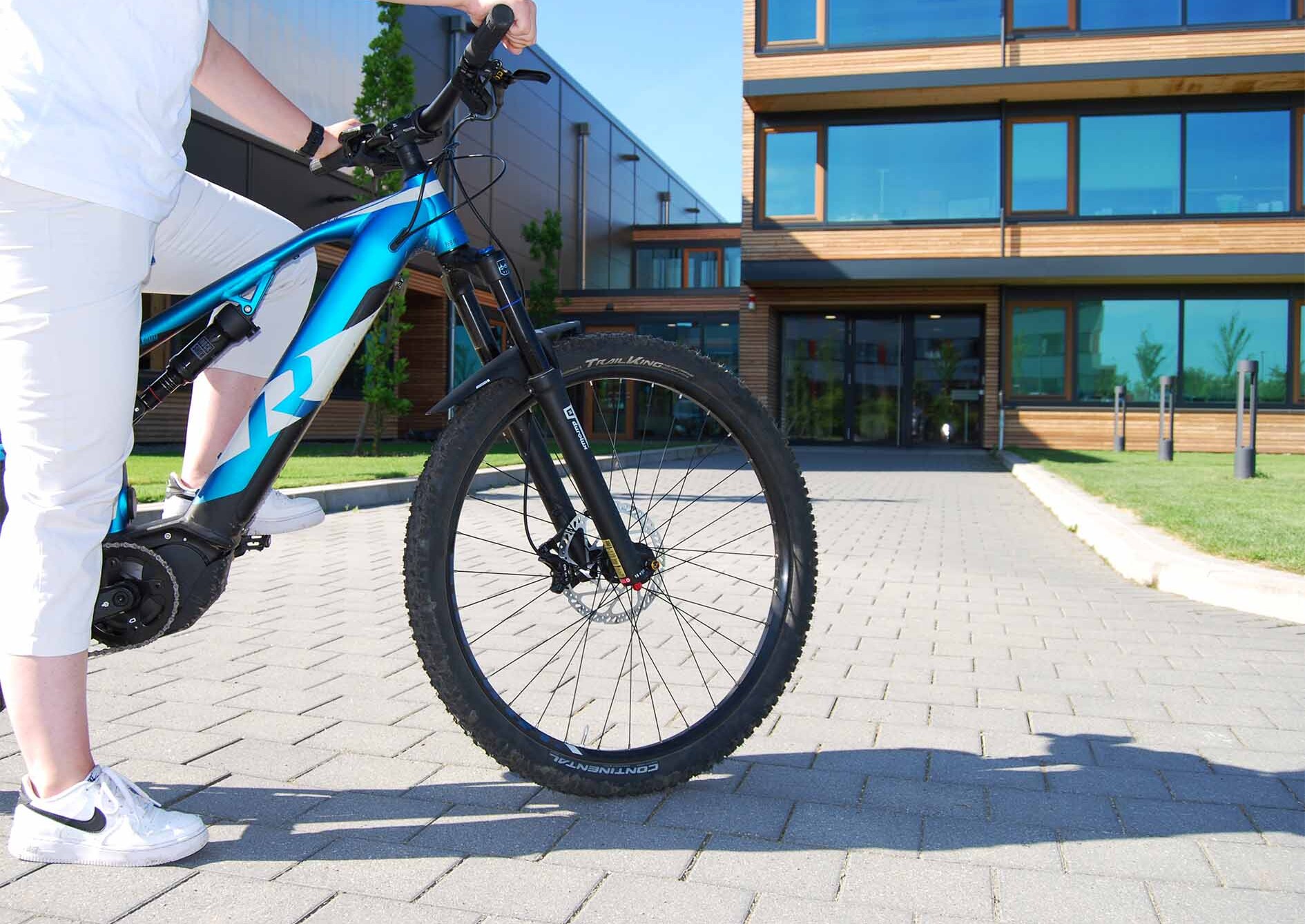 Person in weißer Kleidung auf einem blauen Fahrrad, steht auf einem gepflasterten Weg vor einem modernen Gebäude mit Holzfassade und großen Fenstern.