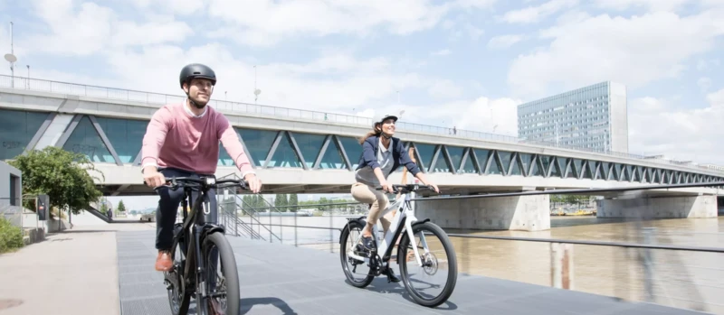 Zwei Personen mit Helmen fahren lächelnd auf E-Bikes entlang eines Flusses, im Hintergrund eine moderne Brücke.