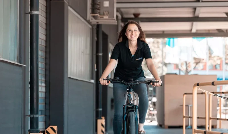 Eine lächelnde Frau fährt auf einem Fahrrad durch einen überdachten Bereich, gekleidet in ein schwarzes Poloshirt und Jeans. Der Hintergrund zeigt eine industrielle Umgebung mit grauen Wänden und Geländern.