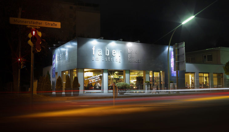 Eine nächtliche Aufnahme eines Restaurants mit der leuchtenden Aufschrift "Faber am Ring" an der Fassade. Vor dem Gebäude sind Lichter von vorbeifahrenden Autos verwischt zu sehen. Eine Straßenampel und ein Straßenschild mit der Aufschrift "Münnersdörfer Straße" befinden sich links im Bild.