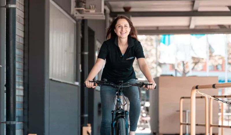 Eine lächelnde Frau fährt auf einem Fahrrad durch einen überdachten Bereich, gekleidet in ein schwarzes Poloshirt und Jeans. Der Hintergrund zeigt eine industrielle Umgebung mit grauen Wänden und Geländern.