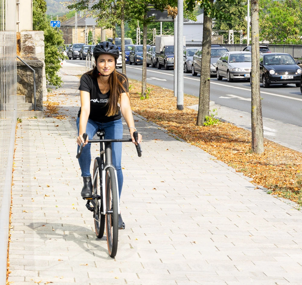 Eine Frau fährt auf einem Fahrrad auf einem breiten Gehweg, trägt einen Helm und ist in Freizeitkleidung unterwegs. Im Hintergrund verläuft eine Straße mit vielen Autos im Verkehr.