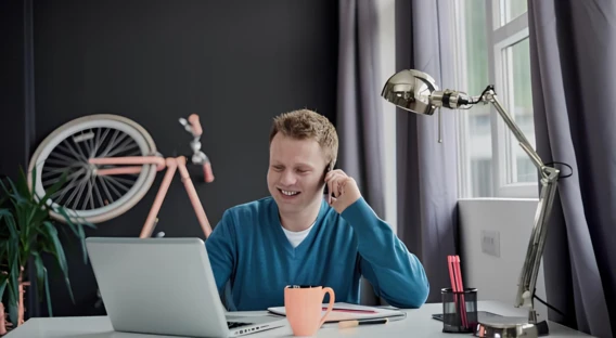 Ein Mann sitzt lächelnd an einem Schreibtisch mit Laptop, Tasse und Lampe, während ein Fahrrad im Hintergrund an der Wand lehnt.