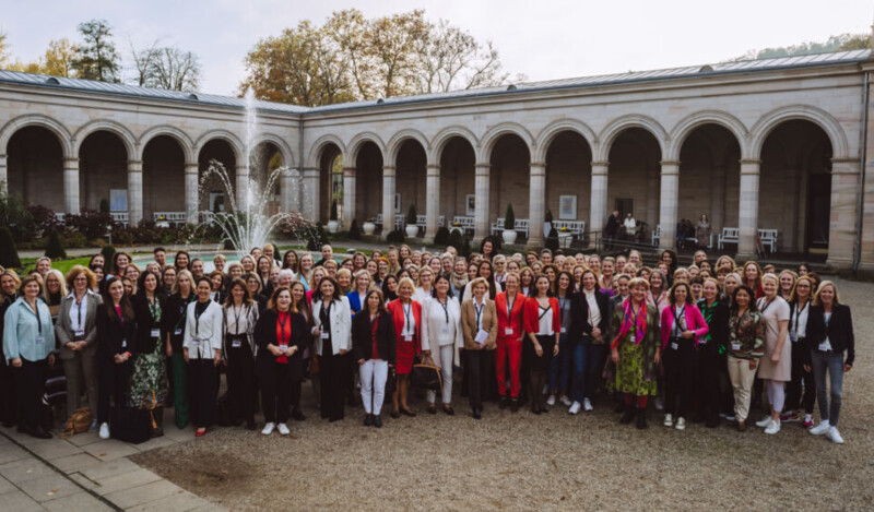 Eine große Gruppe von Menschen, hauptsächlich Frauen, steht vor einem historischen Gebäude mit einer Arkadenreihe und einem Brunnen in der Mitte. Die Gruppe posiert lächelnd für ein Gruppenfoto, umgeben von einer gepflegten Gartenanlage.