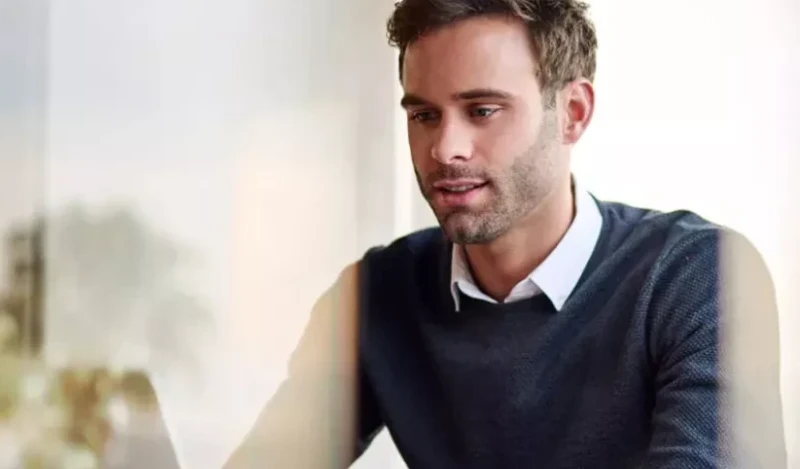 Ein junger Mann mit kurzem braunem Haar sitzt konzentriert an einem Laptop. Er trägt einen dunkelblauen Pullover über einem weißen Hemd. Die Umgebung ist hell und verschwommen, möglicherweise ein modernes Büro oder ein Zuhause mit natürlichem Lichteinfall.