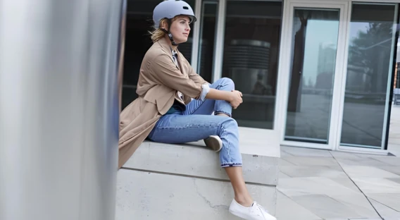 Eine Frau mit Fahrradhelm sitzt entspannt auf einer Steinmauer in einem urbanen Umfeld. Sie trägt eine helle Jeans, weiße Sneaker und einen beigen Mantel und schaut gedankenverloren zur Seite.