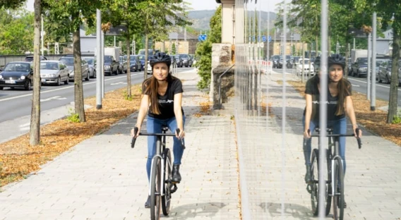 Eine Frau mit Helm sitzt lächelnd auf einem Fahrrad, umgeben von einem modernen städtischen Umfeld mit Pflanzen im Vordergrund.