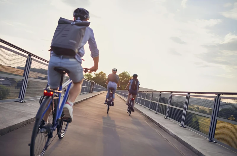 Drei Personen fahren bei Sonnenuntergang auf einem Radweg über eine Brücke. Alle tragen Helme und Rucksäcke, während sie entspannt nebeneinander herfahren.