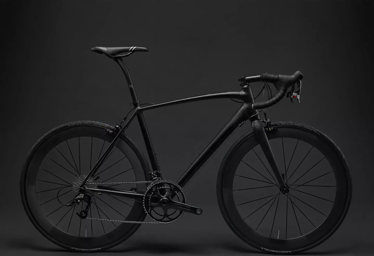 Ein elegantes, mattschwarzes Rennrad vor einem dunklen Hintergrund. Das Fahrrad hat dünne Reifen, aerodynamische Felgen und einen gebogenen Rennlenker, wodurch es modern und minimalistisch wirkt.