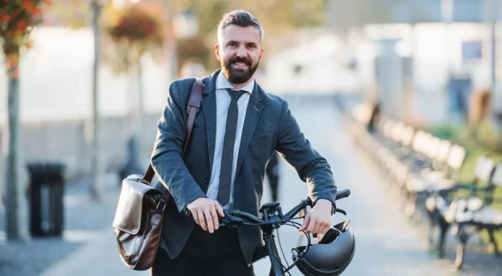 Ein lächelnder Mann in Businesskleidung, mit Sakko und Krawatte, steht draußen neben einem Fahrrad. Er trägt eine Umhängetasche und hält einen Fahrradhelm in der Hand, während die herbstliche Umgebung und eine Allee mit Bänken im Hintergrund sichtbar sind.