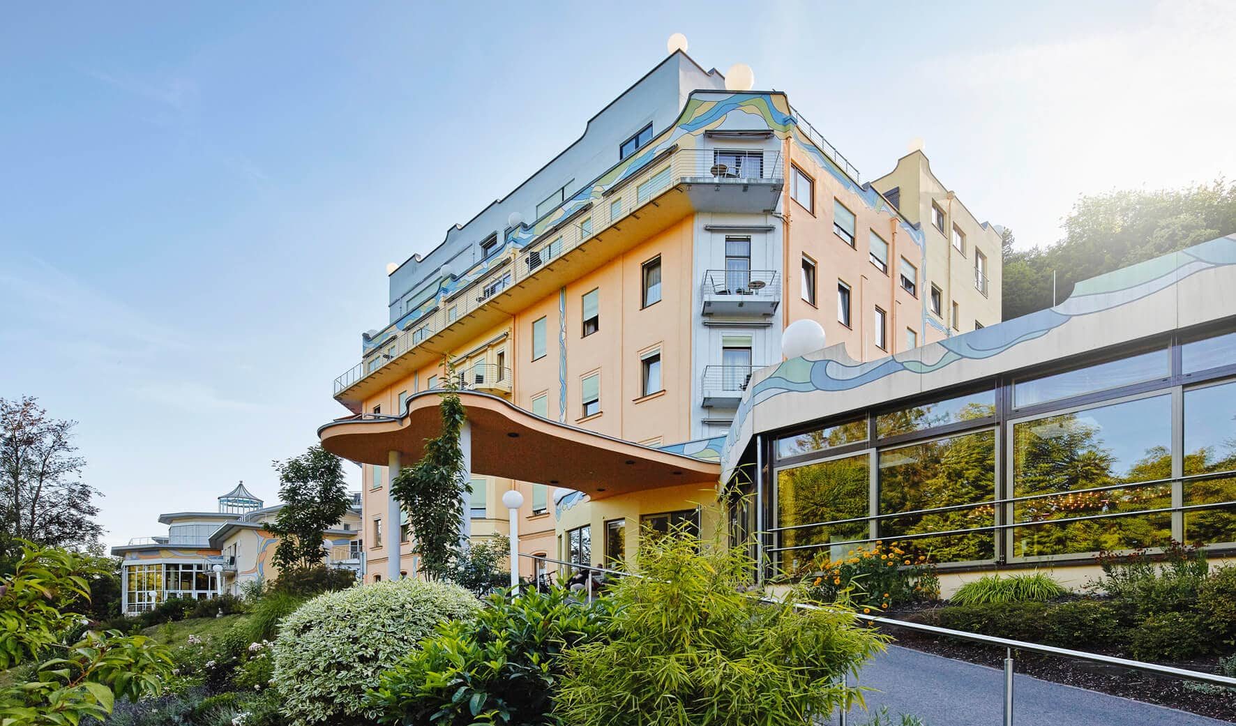 Ein mehrstöckiges, helles Gebäude mit dekorativen Wellenmustern an der Fassade. Vor dem Gebäude befindet sich ein gepflegter Garten mit üppigen Pflanzen. Der Himmel ist klar, und die Szene wirkt ruhig und einladend.