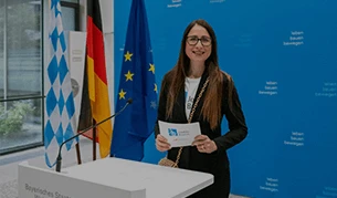 Eine Frau mit langen braunen Haaren steht an einem Rednerpult und hält eine Karte in den Händen. Im Hintergrund sind die Flaggen von Deutschland, der Europäischen Union und Bayern zu sehen, sowie eine blaue Wand mit weißer Aufschrift.