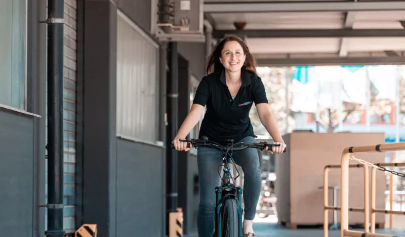 Eine Frau in einem schwarzen T-Shirt fährt lächelnd auf einem Fahrrad durch einen überdachten Bereich.