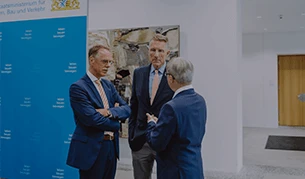 Drei Männer in Anzügen stehen zusammen und unterhalten sich vor einer blauen Wand mit dem Logo des Bayerischen Staatsministeriums für Wohnen, Bau und Verkehr.