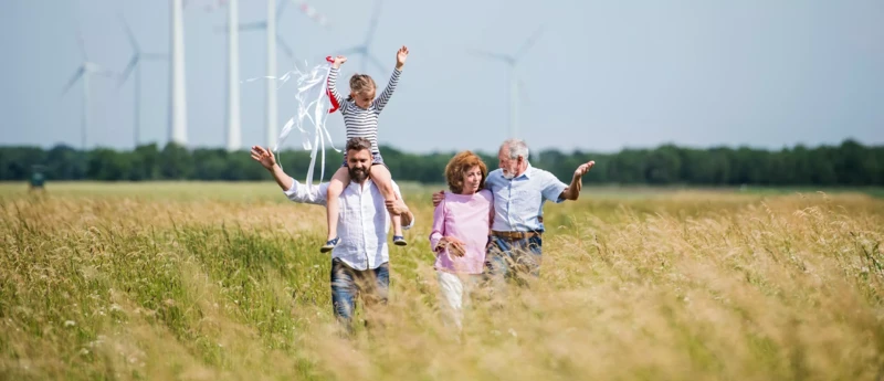 Eine Familie spaziert durch ein Feld; ein Mann trägt ein Kind auf den Schultern, im Hintergrund sind Windräder zu sehen.