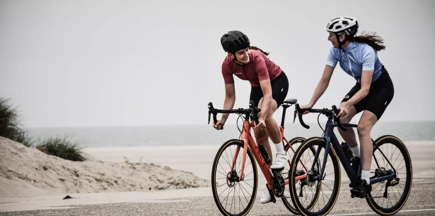 Zwei Frauen fahren Rennräder auf einem sandigen Weg nahe der Küste. Beide tragen Fahrradhelme und sportliche Kleidung. Die Frau links fährt ein orangefarbenes Fahrrad, die Frau rechts ein blaues. Sie lächeln einander an, während sie nebeneinander fahren.